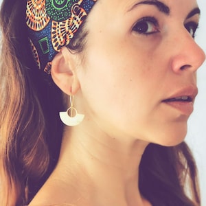 Może przedstawiać: Kobieta z długimi brązowymi włosami nosi opaskę na głowę z kolorowym geometrycznym wzorem. Ma na sobie złote kolczyki w kształcie białego półksiężyca.