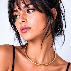 Könnte beinhalten: Eine glatte, massive goldfarbene Metall-Halskette im Choker-Stil mit abgerundetem Profil. Die Halskette liegt flach am Halsansatz an und zeichnet sich durch ein nahtloses, kreisförmiges Design mit einer gleichmäßig glänzenden Oberfläche aus.