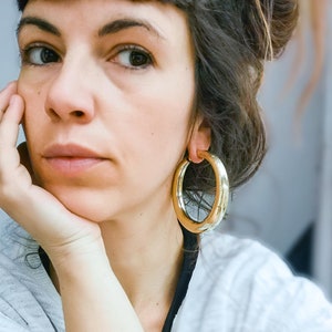 Può includere: Una donna con capelli castano scuro che indossa una camicia grigia chiaro e grandi orecchini a cerchio dorati.