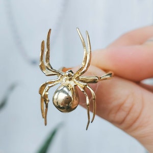 Può includere: Un anello a forma di ragno dorato con un design dettagliato. Il ragno è posizionato con le zampe distese.