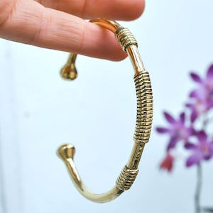 Könnte beinhalten: Ein goldfarbenes Armreif-Armband mit einem strukturierten, spiralförmigen Design in der Mitte. Die offenen Enden haben abgerundete, knollenförmige Spitzen. Das Armband wird vor einem Hintergrund aus lila Blumen und einem hellblauen Hintergrund präsentiert.