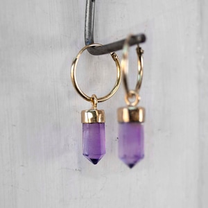 Op de afbeelding: Een paar gouden creolen met paarse amethist kristallen hangers.