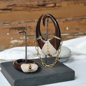 May include: A pair of wooden ear gauges with a chain and decorative beads. The gauges are displayed on a black stand.