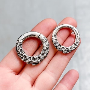 May include: Two silver-colored, textured hoop earrings. The earrings have a unique, organic design with a rough, pitted surface. The hoops are of different sizes, with a hinged clasp for closure. Jewelry.