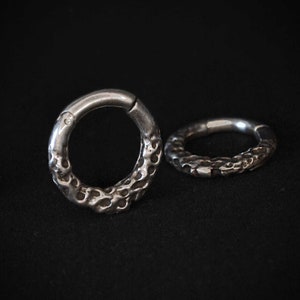 May include: Two silver-toned circular hoop rings featuring a textured, pitted surface on the outer edge. Each ring has a smooth, plain segment near the hinge mechanism, contrasting with the irregular, crater-like detailing on the rest of the band.