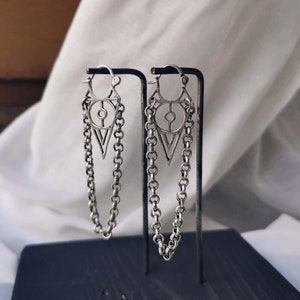May include: A pair of silver earrings with a geometric design and a chain dangle. The earrings are displayed on a black stand.