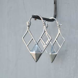 May include: Silver hoop earrings with a geometric diamond-shaped design. The earrings have a pointed bottom and a wire hoop.