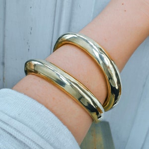 Könnte beinhalten: Zwei goldfarbene, klobige Armreifen werden an einem Arm getragen. Die Armbänder haben eine glatte, polierte Oberfläche und ein abgerundetes, röhrenförmiges Design. Der Arm ist mit einem weißen Langarmshirt bedeckt. Der Hintergrund ist eine verwitterte, weiße Holzoberfläche.