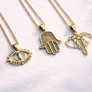 May include: Three gold-coloured pendant necklaces. The pendants feature an eye, a Hamsa hand, and a geometric elephant. Each necklace has a delicate chain. The jewellery is displayed on a white surface.