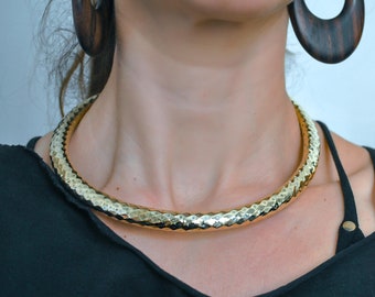 Puede incluir: Un collar tipo gargantilla de metal dorado con una superficie martillada y texturizada que refleja la luz. El collar se lleva con grandes pendientes de aro ovalados de madera de color marrón oscuro. Las joyas se combinan con un top negro de tirantes finos.