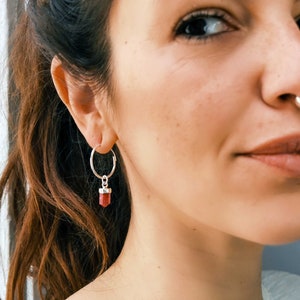 Peut inclure: Gros plan sur l'oreille d'une personne avec une boucle d'oreille en argent en forme de cercle avec une pierre précieuse rouge pendante. La boucle d'oreille est de conception simple avec un petit cercle rond et une seule pierre précieuse rouge qui pend.