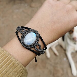 以下が含まれることがあります： 大きな楕円形のムーンストーンがついた黒いマクラメブレスレット。ブレスレットは黒い紐で結ばれており、端に結び目があります。