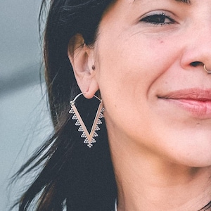 Puede incluir: Una mujer con el pelo oscuro y un piercing en la nariz lleva un par de pendientes triangulares de oro con un diseño de cuentas.