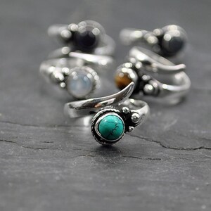 Op de afbeelding: Een set zilveren ringen met verschillende gekleurde edelstenen. De ringen hebben een uniek ontwerp met een gebogen band en een klein, puntig accent. De edelstenen zijn turquoise, maansteen, tijgeroog en zwarte onyx.