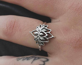 Puede incluir: Un anillo de plata con un delicado diseño de flor de loto. El anillo se lleva en el dedo medio de una mano.