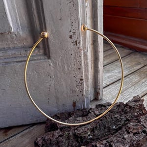 May include: A gold-toned choker necklace with round ends. The necklace is lying on a wooden surface.