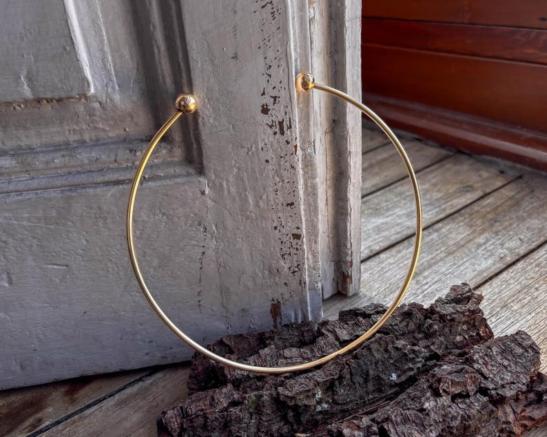 May include: A gold-toned choker necklace with round ends. The necklace is lying on a wooden surface.