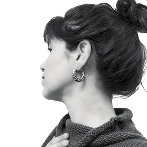 May include: A black and white image of a person wearing a pair of textured silver hoop earrings. The earrings are the focal point, with a detailed, patterned surface. The person is wearing a dark gray sweater.