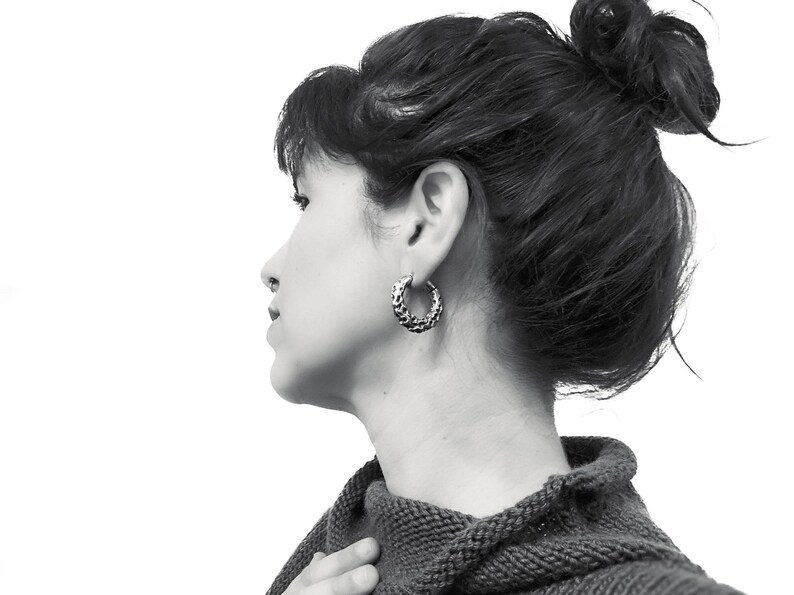 May include: A black and white image of a person wearing a pair of textured silver hoop earrings. The earrings are the focal point, with a detailed, patterned surface. The person is wearing a dark gray sweater.