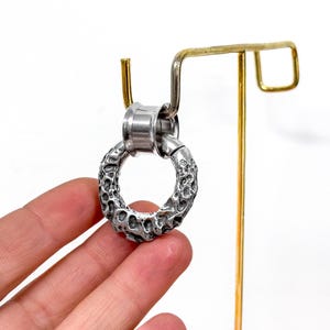 May include: A silver-toned circular ear gauge with a textured, cratered surface. The jewelry piece features a smooth, polished cylindrical tunnel insert at the top, designed for insertion into a stretched earlobe piercing.