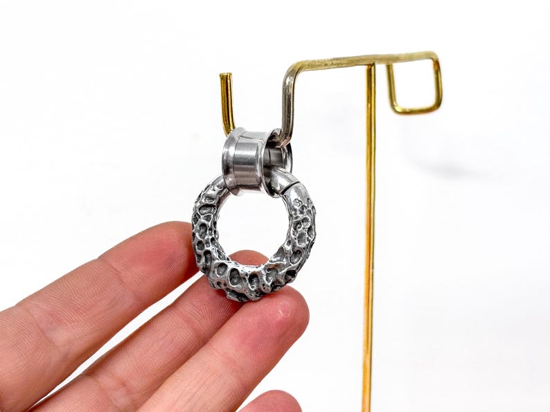 May include: A silver-toned circular ear gauge with a textured, cratered surface. The jewelry piece features a smooth, polished cylindrical tunnel insert at the top, designed for insertion into a stretched earlobe piercing.