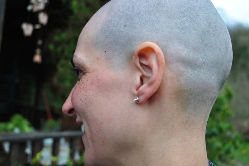 May include: A close-up of a person's head with a shaved head. The person is looking to the right and has a small silver earring in their right ear.