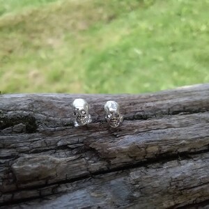 May include: Two silver skull earrings on a wooden surface.