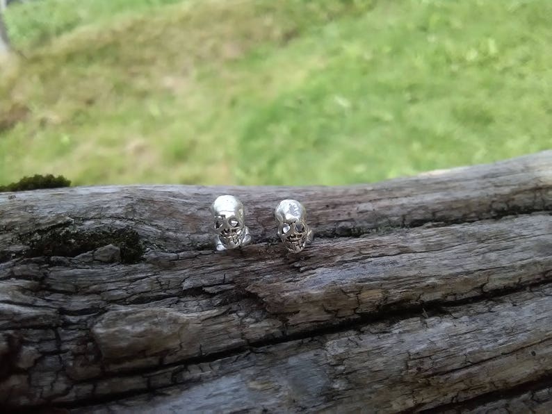 May include: Two silver skull earrings on a wooden surface.