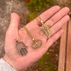 May include: Three gold-coloured pendant necklaces. One features a Hamsa hand design, another an eye, and the third a geometric elephant. The necklaces are displayed on an open hand against a blurred outdoor background.
