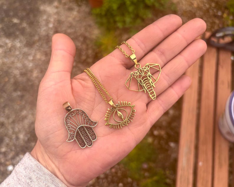 May include: Three gold-coloured pendant necklaces. One features a Hamsa hand design, another an eye, and the third a geometric elephant. The necklaces are displayed on an open hand against a blurred outdoor background.