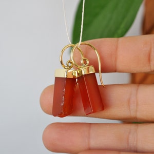Puede incluir: Un par de pendientes de oro con gotas de piedra de cornalina. Las piedras son de color rojo anaranjado oscuro y de forma rectangular.