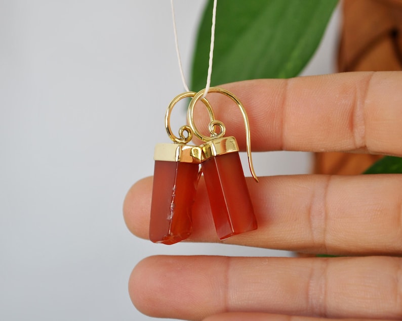 Puede incluir: Un par de pendientes de oro con gotas de piedra de cornalina. Las piedras son de color rojo anaranjado oscuro y de forma rectangular.