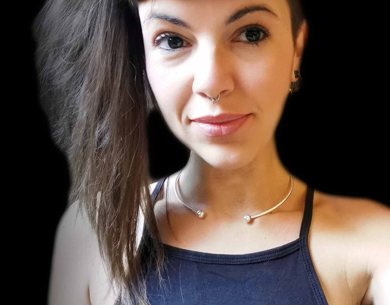 May include: A woman with long brown hair wearing a black tank top and a silver choker necklace with two small round beads.
