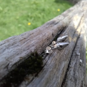 May include: A pair of silver bullet-shaped earrings with a detailed design on the front. The earrings are on a wooden surface.