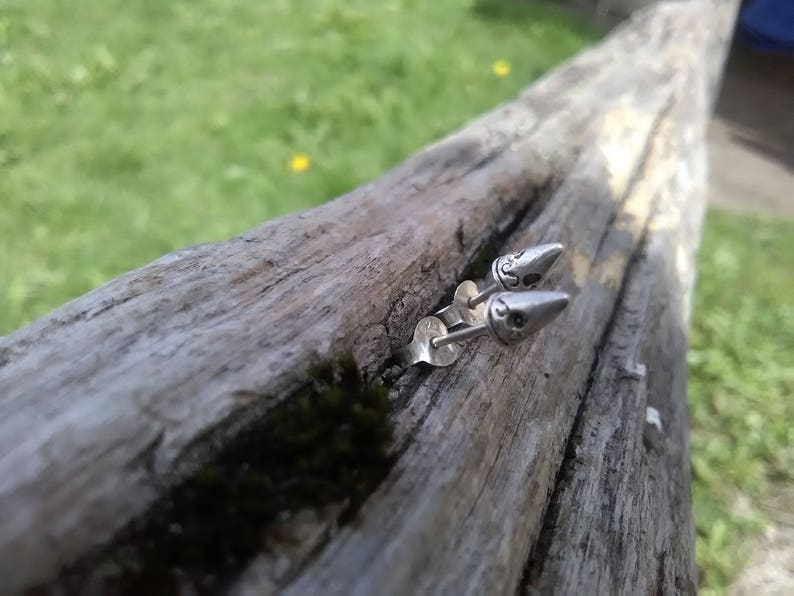 May include: A pair of silver bullet-shaped earrings with a detailed design on the front. The earrings are on a wooden surface.