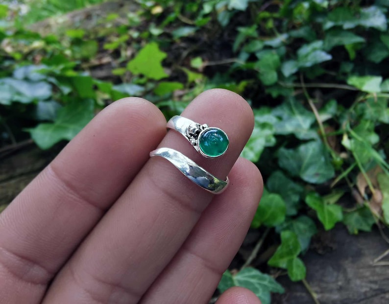 Op de afbeelding: Een zilveren ring met een groene edelsteen gezet in een decoratieve zilveren setting. De ring is ontworpen om om de vinger te wikkelen.