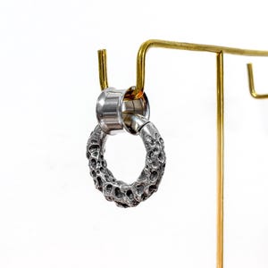 May include: A silver-colored earring with a textured, porous design. The earring is a circular shape and is attached to a silver-colored post. The earring is displayed on a gold-colored earring stand against a white background.