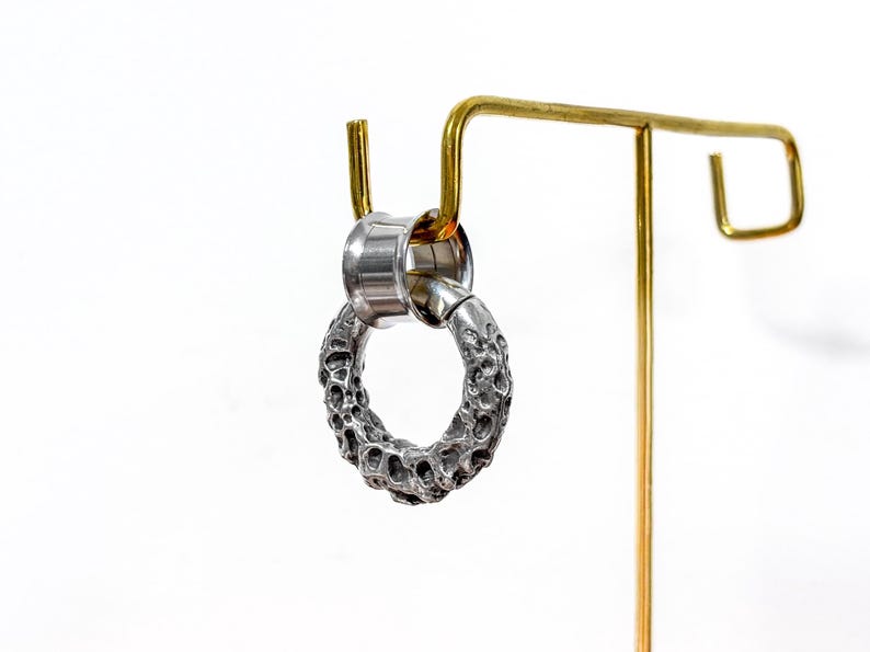 May include: A silver-colored earring with a textured, porous design. The earring is a circular shape and is attached to a silver-colored post. The earring is displayed on a gold-colored earring stand against a white background.