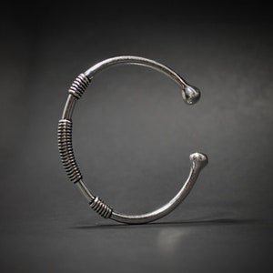 May include: A silver cuff bracelet with a distinctive design. The bracelet has a coiled wire section in the middle and rounded ends. The metal has a polished finish, and the bracelet is set against a dark background.