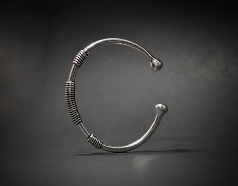 May include: A silver cuff bracelet with a distinctive design. The bracelet has a coiled wire section in the middle and rounded ends. The metal has a polished finish, and the bracelet is set against a dark background.