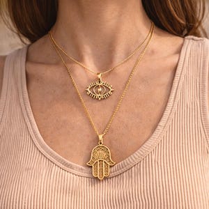 May include: Two gold necklaces. The top necklace has an eye-shaped pendant, and the bottom one features a Hamsa hand pendant. Both pendants are suspended from fine chains. The necklaces are shown against a neutral backdrop.