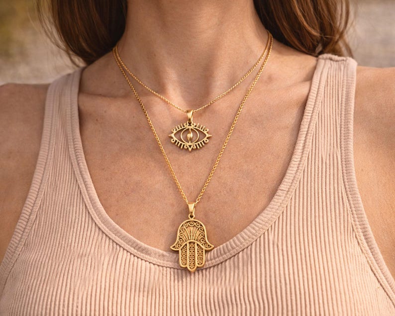 May include: Two gold necklaces. The top necklace has an eye-shaped pendant, and the bottom one features a Hamsa hand pendant. Both pendants are suspended from fine chains. The necklaces are shown against a neutral backdrop.