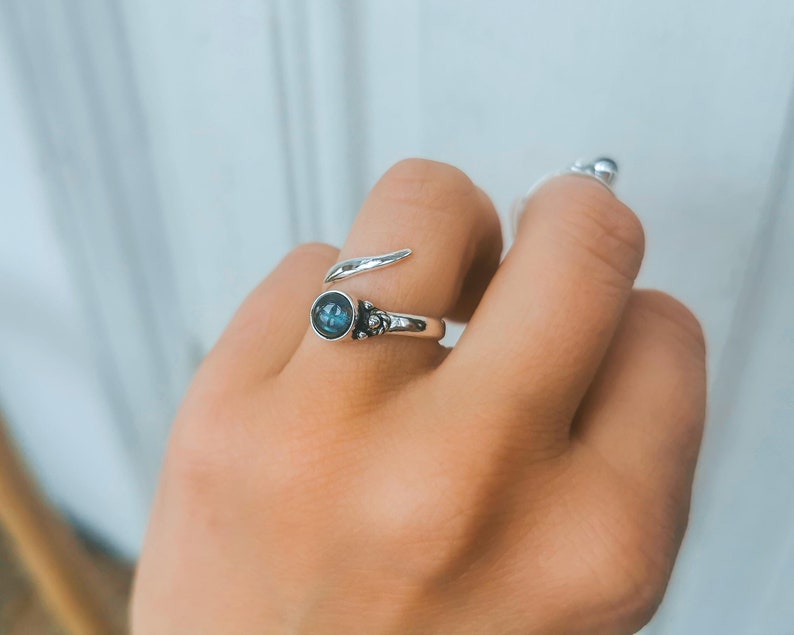 Op de afbeelding: Een zilveren ring met een blauwe edelsteen en een gedraaide band. De ring zit aan een vinger.