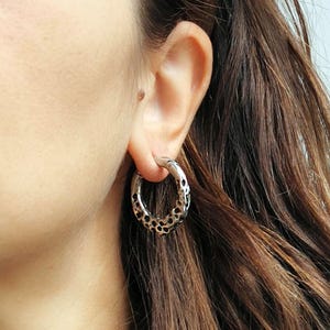 May include: Silver-toned hoop earring with a textured, dotted pattern. The earring is circular and has a polished finish. The earring is worn on the ear of a person with brown hair. The jewelry is a fashion accessory.