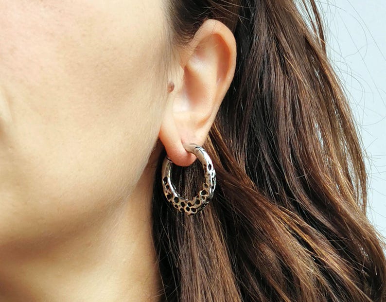 May include: Silver-toned hoop earring with a textured, dotted pattern. The earring is circular and has a polished finish. The earring is worn on the ear of a person with brown hair. The jewelry is a fashion accessory.
