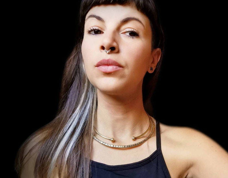 May include: A woman with dark hair and a silver streak in her hair is wearing a black tank top and a gold choker necklace. She has a nose ring and a small earring in her right ear.
