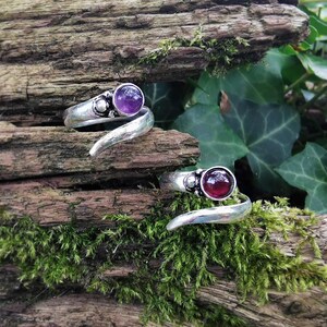 Op de afbeelding: Twee zilveren ringen met een zwierig ontwerp. De ene ring heeft een paarse edelsteen en de andere een rode edelsteen.