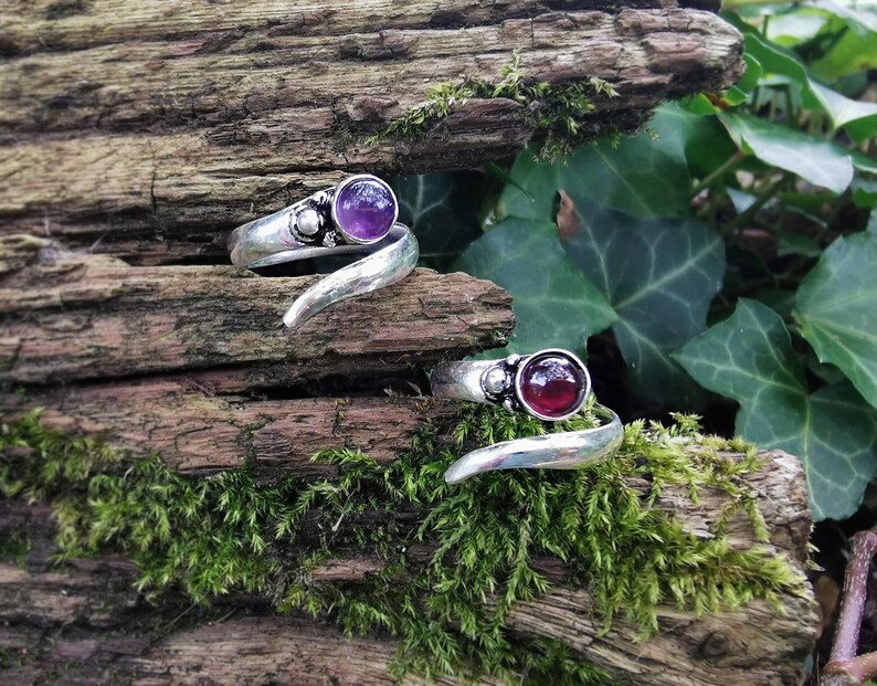 Op de afbeelding: Twee zilveren ringen met een zwierig ontwerp. De ene ring heeft een paarse edelsteen en de andere een rode edelsteen.