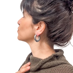 May include: Silver hoop earrings with a textured, braided design. The earrings are worn by a person with dark hair and a brown sweater. The background is plain white.