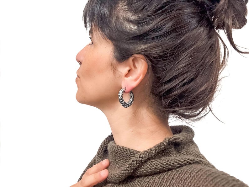 May include: Silver hoop earrings with a textured, braided design. The earrings are worn by a person with dark hair and a brown sweater. The background is plain white.